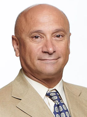 The image shows a man with short gray hair smiling at the camera. He has a light tan complexion and is wearing a brown jacket over a blue shirt, paired with a patterned tie.