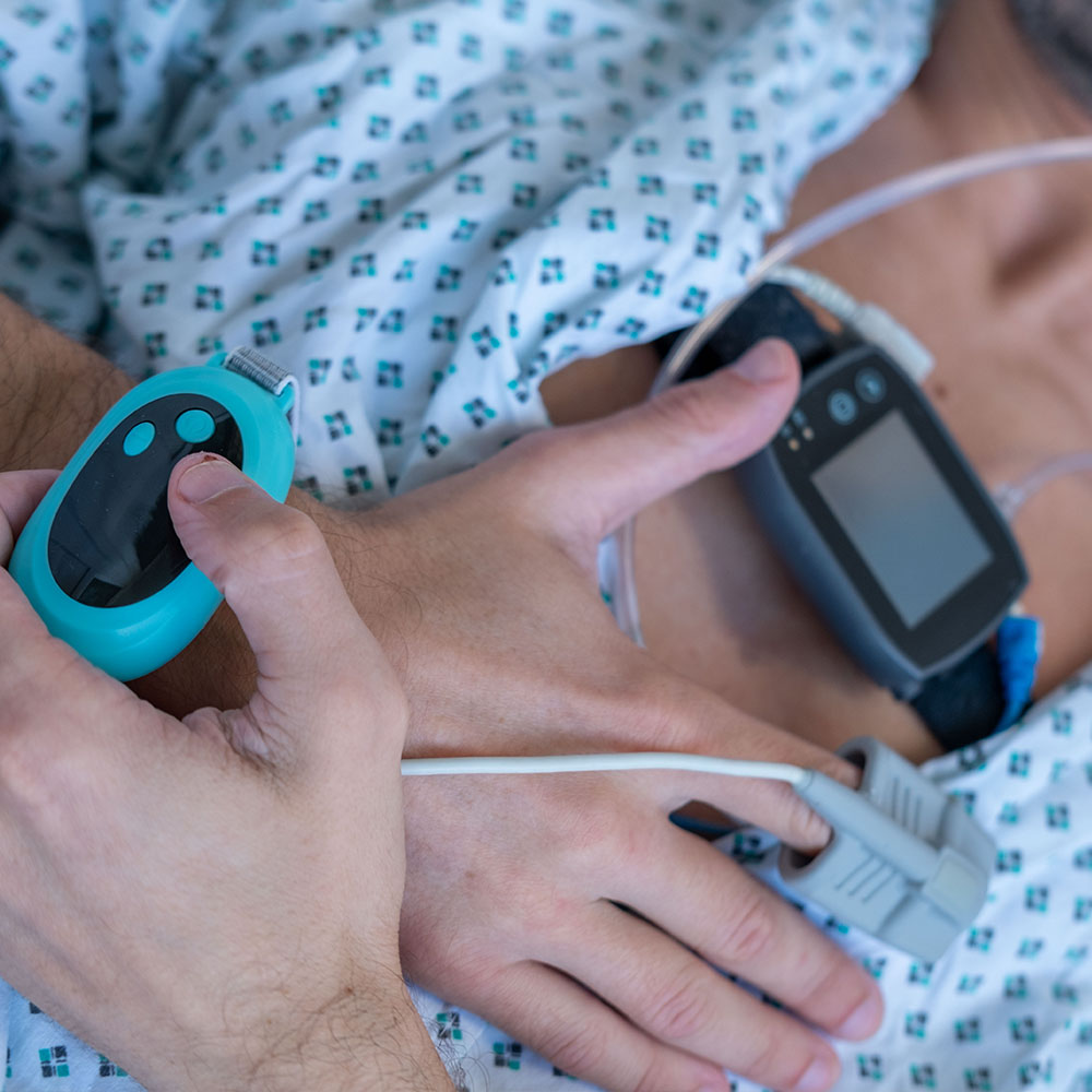 The image shows a person lying in a hospital bed with medical equipment attached to their body, including a heart monitor and oxygen supply, being attended to by a healthcare professional who is holding an electronic device and wearing gloves.