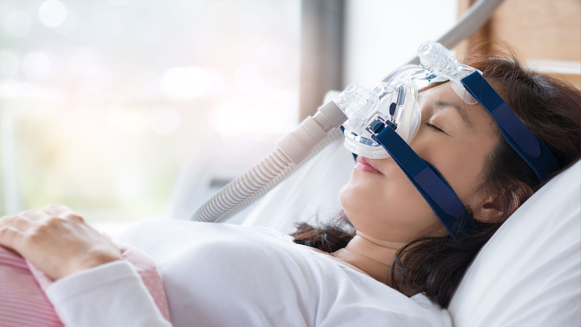 The image shows a person lying down with a medical device attached to their face, likely for oxygen therapy, in a room with natural light coming through a window.