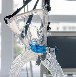 The image shows a mechanical ventilator with its hose connected to a mask, set up against a white background.