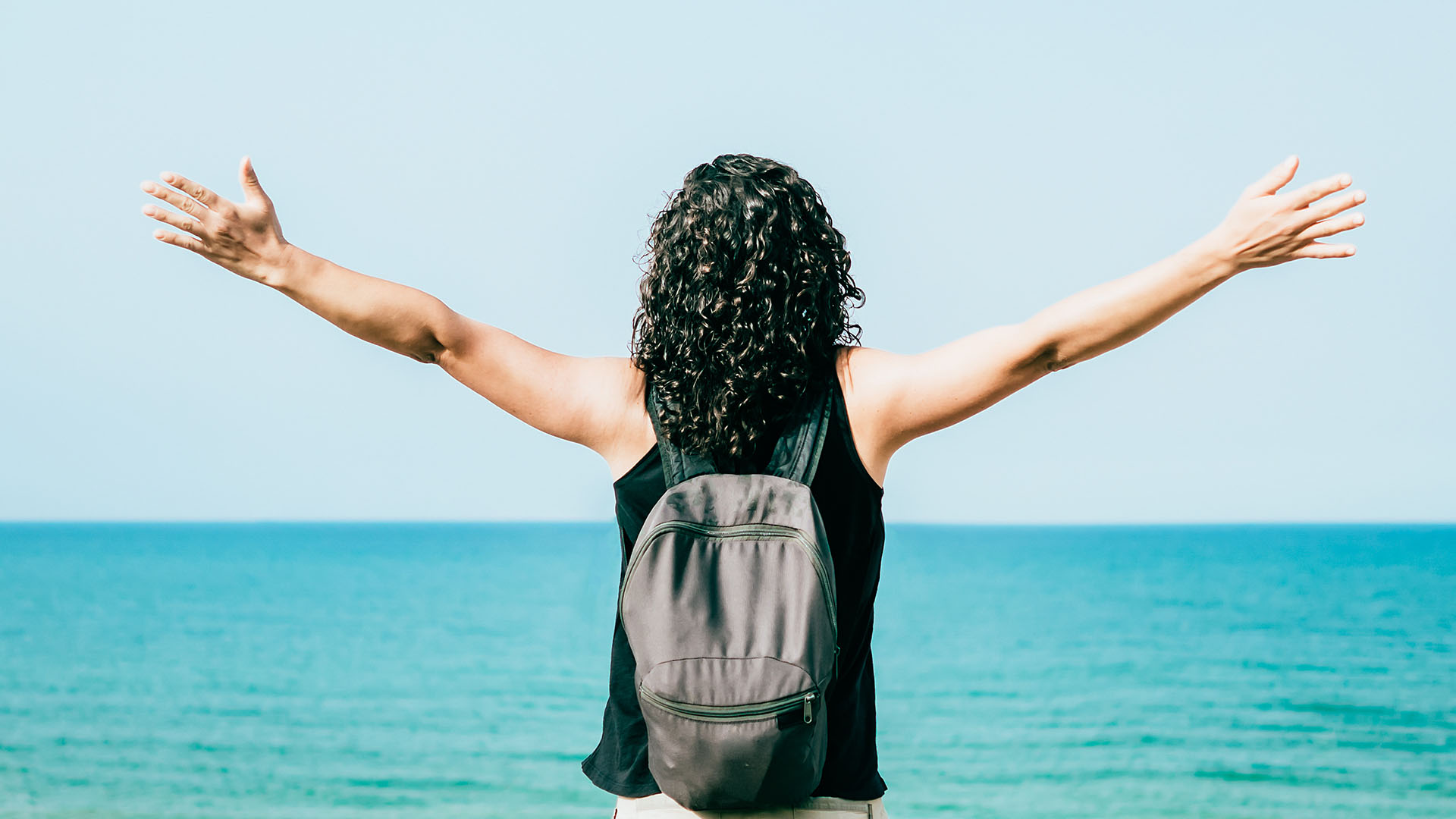 A person with their arms raised high, standing on a beach against a backdrop of the ocean.